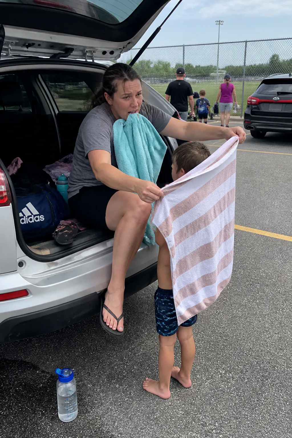 A parent juggling towels at the back of an open trunk in a parking lot